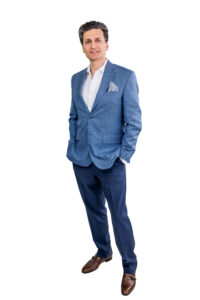 Full-length portrait of a man in a blue textured suit, white shirt, pocket square, standing with hands in his pockets against a white background.
