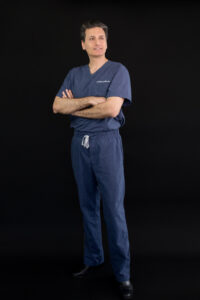 Full-length portrait of a man in a dark blue surgical scrubs, looking to left,, standing with arms folded against a dark background.